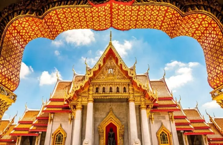 Temple bouddhiste richement décoré en Thaïlande, encadré par une arche traditionnelle dorée.