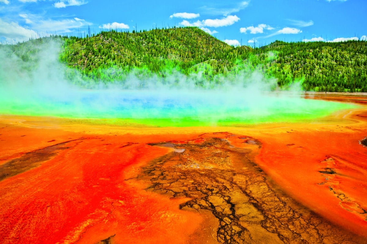 Beautiful cerulean geyser surrounded by colorful layers of bacteria, against cloudy blue sky.