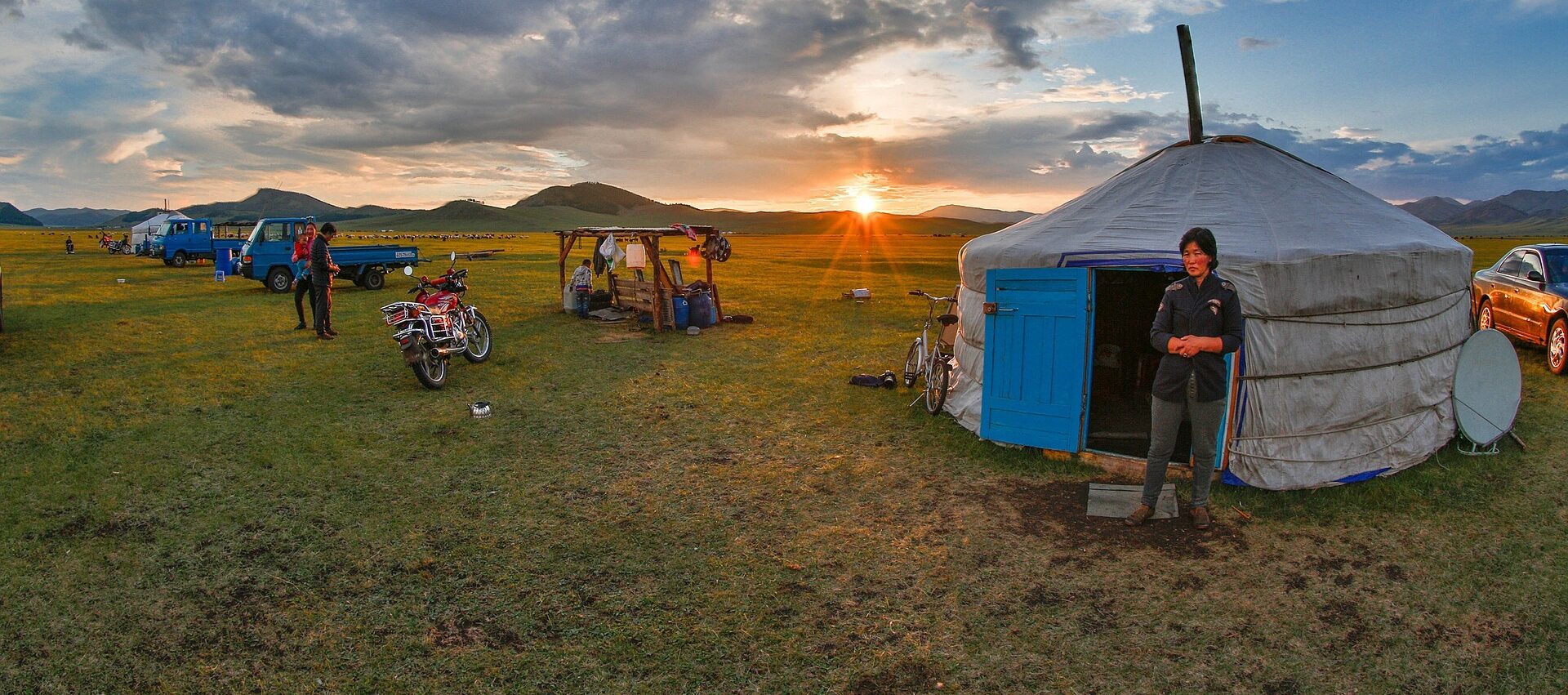 Campement nomade au coucher du soleil en Mongolie avec yourte traditionnelle et véhicules, reflétant la vie nomade moderne.