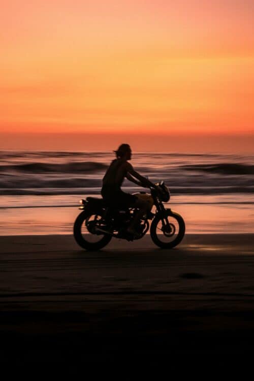 Un motard silhouette sur une plage de Bali au coucher du soleil avec des couleurs oranges et roses dans le ciel.