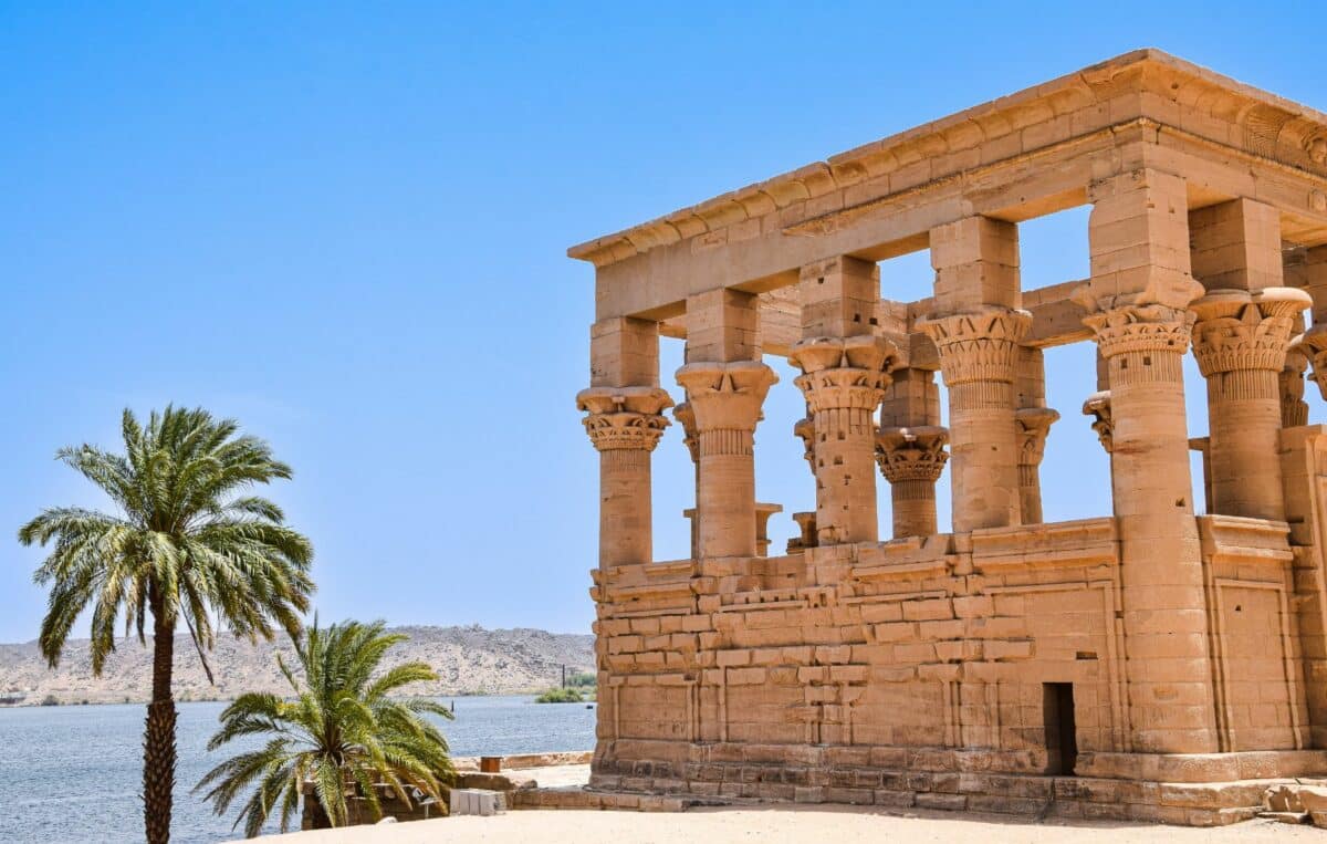 Temple de Philae au bord du Nil avec palmiers et ciel bleu.