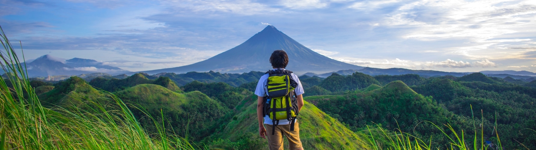 Voyageur contemplant un volcan au lever du soleil, entouré de collines verdoyantes.