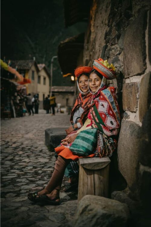 Deux jeunes filles quechuas en tenue traditionnelle assises dans une ruelle pavée d’Ollantaytambo.