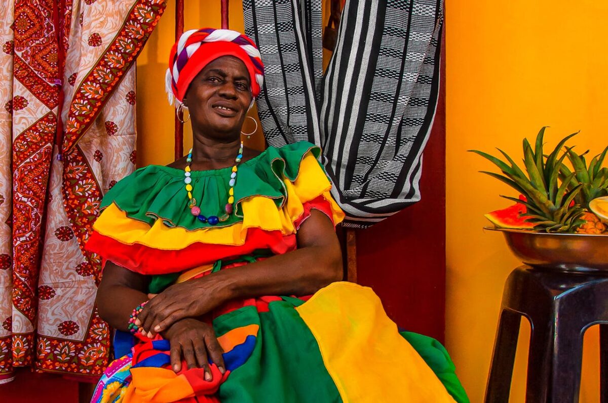 Femme en robe traditionnelle assise devant des étoffes colorées.